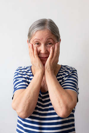Elderly woman, gray-haired lady of the 60s in a striped white and blue dress shocked, surprised and amazed expression with hands on her face on a white background.の写真素材