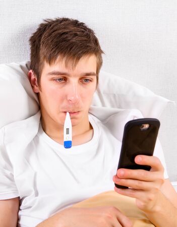 Young Man with a Phone and a Thermometer in the Bed at the Homeの写真素材
