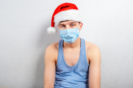 Sad Young Man in Santa Hat and Flu Mask by the Wall in the Roomの写真素材