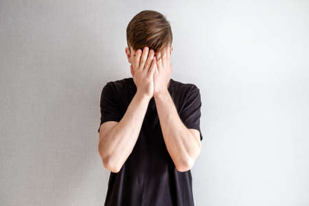 Sad Young Man close his Face with a Hands by the White Wall in the Room