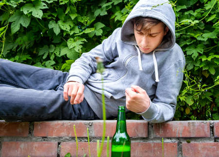 Sad Young Man in a Hoodie with a Beer Bottle on the Sidewalk of the City Streetの写真素材