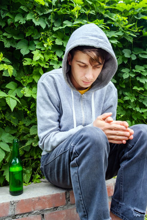 Sad Young Man in a Hoodie with a Beer Bottle on the Sidewalk of the City Streetの写真素材