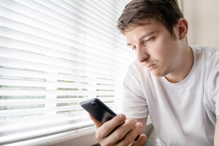 Sad Young Man with a Phone by the Window in the Roomの写真素材