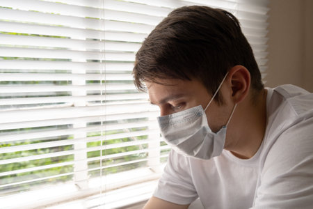 Sad Young Man in a Surgical Mask by the Window in the Roomの写真素材
