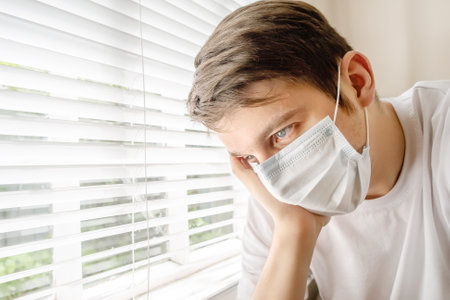 Sad Young Man in a Surgical Mask by the Window in the Roomの写真素材