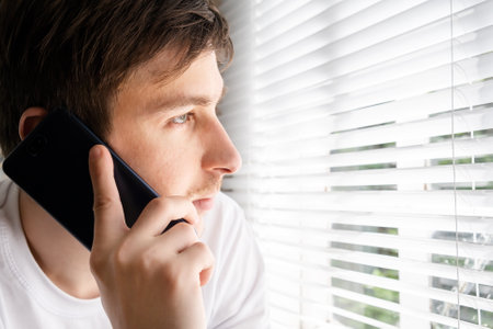 Sad Young Man with a Phone by the Window in the Roomの写真素材