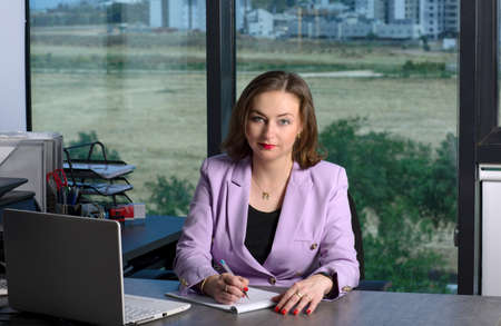 Happy business woman in smart casual clothes sitting at the office table and looks at the camera in...