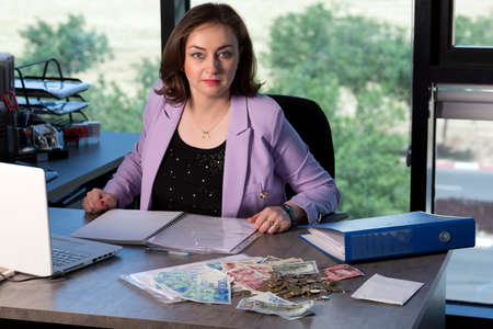 Portrait of happy beautiful brunette working at her office desk with documents, laptop, dollars,...