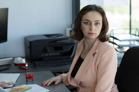Confident business woman in pink suit sitting at the office table. Stylish woman in smart casual clothes sitting and looks at the camera with a serious face. Portrait of beautiful brunette womanの写真素材