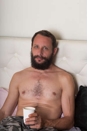 Bearded man drinking coffee from paper cup in bed at home in morning. Rest and people concept. Shirtless man lying in bed with a cup of hot drink. Confident male with nude torso stares at cameraの写真素材