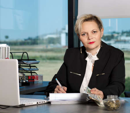 beautiful blonde woman in business suit, smiling at desk behind laptop computer and papers in...