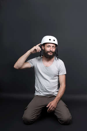 Smiling American handsome bearded man in a bicycle protective helmet standing on his knees. Russian biker sitting on floor on gray background. Active person point index finger on hard hatの写真素材