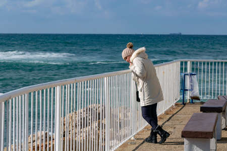 Happy caucasian american woman standing on the beach in a white down jacket and knitting hat with a view of the blue sea and enjoying freedom. Emotional woman walking on white pier against the seaの写真素材