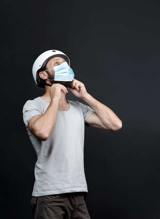 Handsome American bearded man working in package delivery, wears a bike helmet with a surgical mask to be safe during pandemic. Russian biker fastening bicycle helmet on gray background.の写真素材