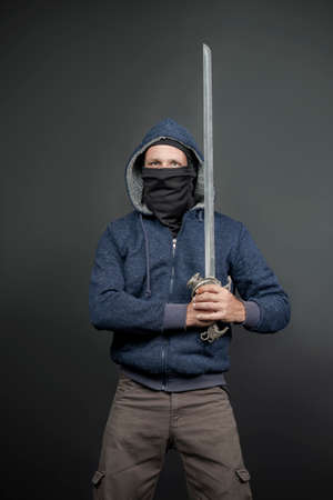 American Brutal man in a blue hoodie and black face mask with a Fantasy Handle Sword, stern look. Portrait of a courageous warrior in jeans and a hood holding a big sword with two hands after a battleの写真素材