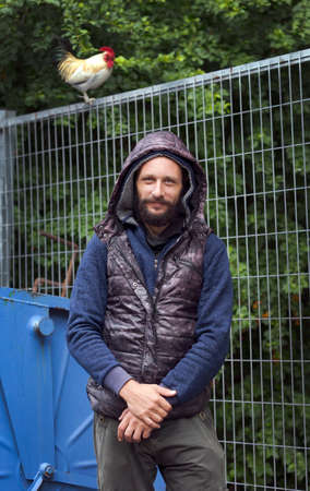 Stylishly american man with a beard dressed in green winter jacket with a hood stands against the background of blurred white rooster perched on fence on the street. Russian man in vest with hoodの写真素材