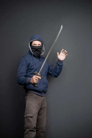 American Brutal man in a blue hoodie and black face mask with a Japanese sword. Portrait of a courageous warrior in jeans and a hood raising and holding his sword up isolated on gray backgroundの写真素材