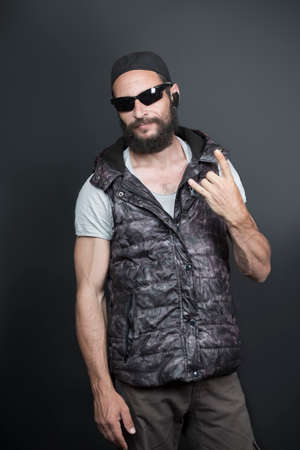 American handsome bearded man wearing a cap, sunglasses, hoody vest Making A Punk Rock Sign. Serious Russian Man with open vest on with hood showing Gesture that rocks or horns. Rock roll signsの写真素材