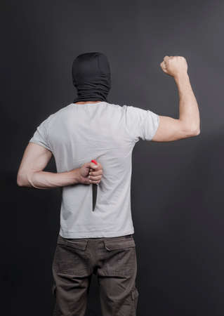 An evil american man holds a kitchen knife as weapon behind his back, waiting for the victim. A maniac and a rapist with a knife in mask on gray background. Poster with space for copy or your textの写真素材