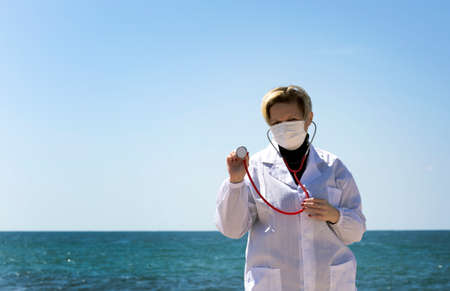 Blond doctor woman of Russian Caucasian appearance Holding A Stethoscope In Hand. Portrait of professional confident American doctor in medical mask and white coat, stethoscope over neck on a beachの写真素材