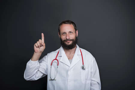 Middle age handsome Caucasian American doctor bearded man wearing doctor uniform and stethoscope on black background smiling with an idea or question pointing finger up with happy face, number oneの写真素材