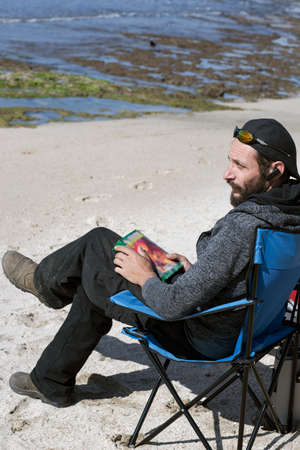 American man relaxing on a book reading on the beach during sunset. Russian Man reading book in deck chair overlooking Sea. handsome Caucasian bearded man wearing sunglasses, cap and hood jacketの写真素材