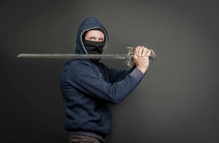 American Brutal man in a blue hoodie and black face mask with a Japanese sword. Portrait of a courageous warrior in jeans and a hood holding a big sword on the shoulder isolated on gray backgroundの写真素材