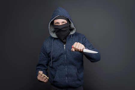 an offender attack with Brass knuckles and knife. Metal brass knuckles in the hand of American hooded man in mask. forbidden weapons in a fight, criminal. caucasian man strike fist with brass knucklesの写真素材