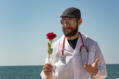 A white happy Bearded Jewish doctor of Caucasian appearance smells a red rose and smiles cheerfully. American Russian man wearing stethoscope, sunglasses, cap, coat against blue sky, holding red roseの写真素材
