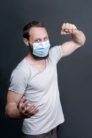 Metal brass knuckles in the hand of a bearded man in a surgical mask. forbidden weapons in a fight, a heavy blow, street thugs. American man showing fist with brass knuckles. Concept of criminalの写真素材