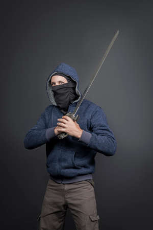 American Brutal man in a blue hoodie and black face mask with a fantasy sword. Portrait of a courageous warrior in jeans and a hood holding a big sword on the shoulder isolated on gray backgroundの写真素材
