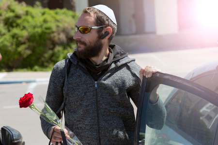 Bearded Charming American Jewish man in white Yarmulke (hat, Kippah, jewish hat) wearing sunglasses and hood jacket getting out of car in town. Brutal Russian Man holding a red rose flower in hand.の写真素材