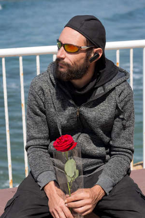 American handsome bearded man wearing sunglasses, cap, hood jacket with rose waiting his woman on the sea beach. Russian Caucasian brutal fashion male looks to side, relaxed pose with natural faceの写真素材