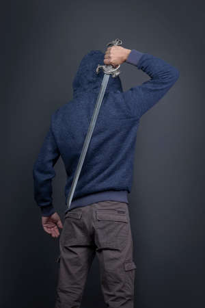 American man in a blue hoodie with a Japanese sword, facing away. Portrait of a courageous warrior in jeans and a hood standing with back towards the camera while holding a sword behind his backの写真素材