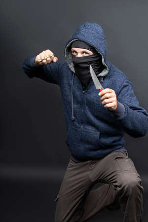 an offender attack with Brass knuckles and knife. Metal brass knuckles in the hand of American hooded man in mask. forbidden weapons in a fight, criminal. caucasian man strike fist with brass knucklesの写真素材