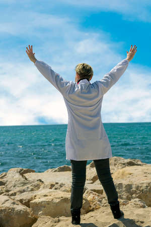 happy Caucasian American Medical woman In Lab Coat enjoying freedom with open hands on sea. Confident female doctor posing on the beach. Portrait Of Doctor walking to the sea, raising hands upの写真素材