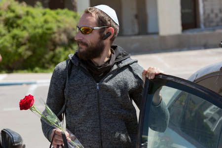 Bearded Charming American Jewish man in white Yarmulke (hat, Kippah, jewish hat) wearing sunglasses and hood jacket getting out of car in town. Brutal Russian Man holding a red rose flower in hand.の写真素材