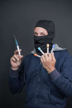 Criminal Caucasian American man in blue hoodie and black face mask holding syringe with toxic acid, preparing to kill enemy. Bad Medicine. Crazy doctor holding syringe with injection. Virus vaccineの写真素材
