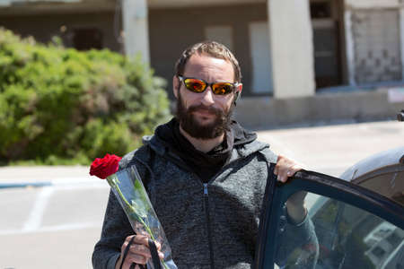 cheerful American handsome bearded man wearing sunglasses and hood jacket getting out of car in town. Brutal Russian Man holding a red rose flower in hand. Romantic, valentines day holiday conceptの写真素材