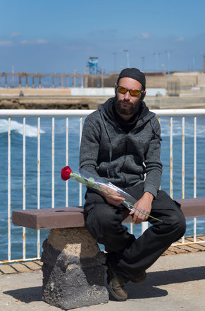 American handsome bearded man wearing sunglasses, cap, hood jacket with rose waiting his woman on the sea beach. Russian Caucasian brutal fashion male sitting on a bench. Romantic date on the beachの写真素材