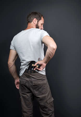 Caucasian American man pulls out a gun tucked in his pant rear view. bearded criminal, illegal, killer holding with Handgun. israeli russian male reaching handgun attached on belt in robbery conceptの写真素材