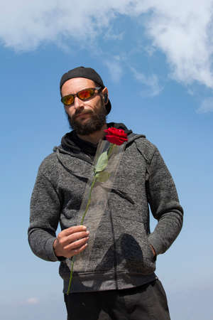 American handsome bearded man wearing sunglasses, cap and hood jacket against blue sky. Portrait of Russian Caucasian brutal dude holding a red rose flower in hand. Romantic and valentines day holidayの写真素材