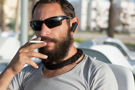 brutal Russian Caucasian beautiful white cool guy smoking wearing sunglasses and gray t-shirt in a cafe. American man with a cigarette in his hand. handsome Jewish man man mustache and beard, close upの写真素材