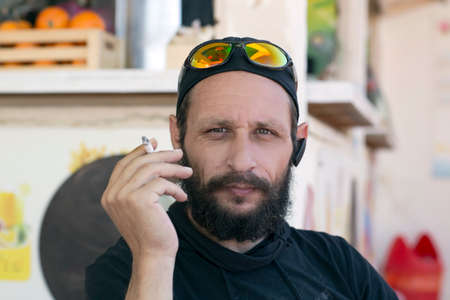 Portrait of a white cool man smoking cigarette wearing sunglasses, cap and hood jacket on the street. Hipster American man smoking in real life. handsome Jewish man bearded man mustache and black hatの写真素材