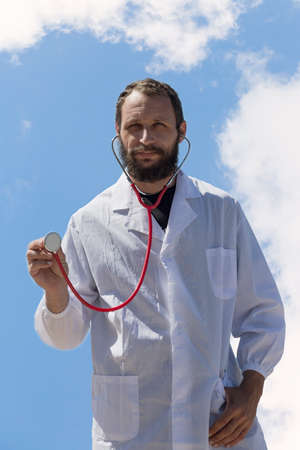 portrait of doctor with stethoscope in ear listen you carefully attentively. American handsome bearded man on blue sky background. Russian Caucasian brutal man in a white coat. Treatment in Israelの写真素材