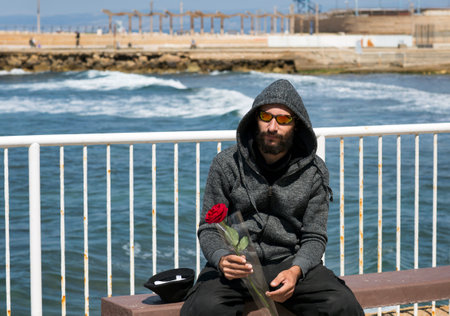 American handsome bearded man wearing sunglasses and hood jacket against blue sea in sunny day. Russian Caucasian brutal dude holding a red rose flower in hand. Romantic and valentines day holidayの写真素材