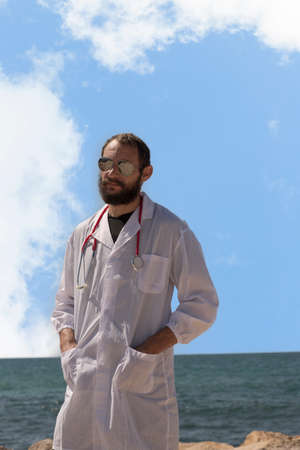 The stethoscope is hanging around the young doctor neck. American handsome bearded man on the sea. Russian Caucasian brutal man in a white coat and sunglasses. Concept of Treatment in Israelの写真素材