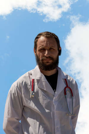 The stethoscope is hanging around the young doctor neck. American handsome bearded man on an isolated blue sky background. Russian Caucasian brutal man in a white coat. Concept of Treatment in Israelの写真素材