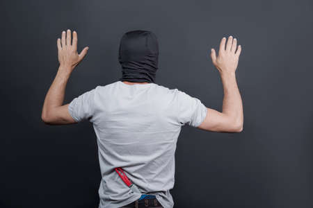 Arrested masked thief with raised arms. The bandit in a black mask, american man holds a kitchen knife as weapon behind his back with hands up. arrest of the criminal, gangster. Back view, Copy spaceの写真素材