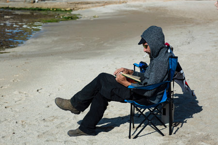American man relaxing on a book reading on the beach during sunset. Russian Man smoking cigarette in deck chair overlooking Sea. handsome Caucasian bearded man wearing sunglasses, cap and hood jacketの写真素材